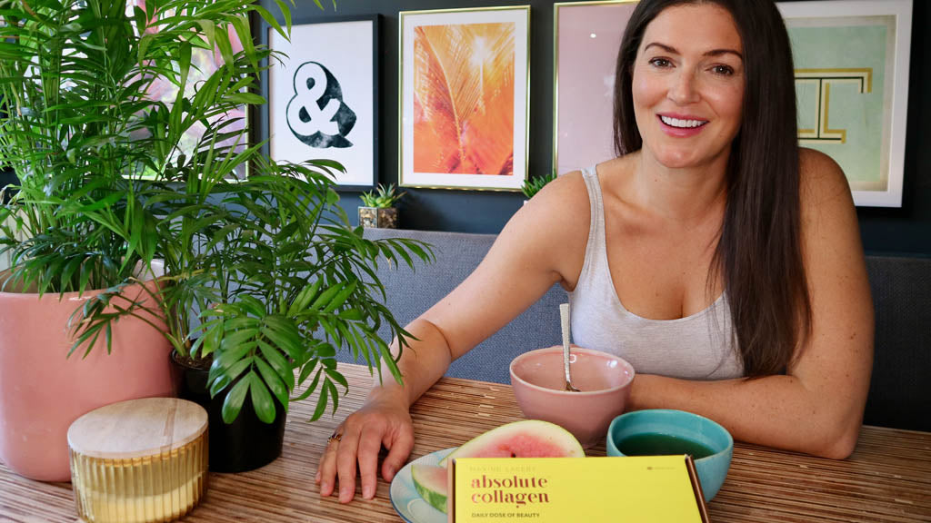 Photo showing a smiling white woman with long brown hair, she is wearing a white vest top, smiling at the camera, and sitting at a table with a box of Absolute Collagen