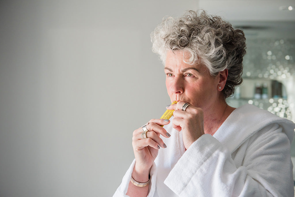 Photo of a white woman with short grey hair wearing a white dressing gown and taking a yellow Absolute Collagen sachet