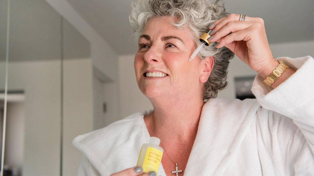 Photo of Maxine Laceby smiling into a mirror and applying Maxerum serum while wearing a white dressing gown